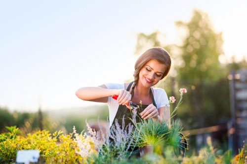 A woman with light brown hair and a bright smile is tending to a diverse garden bed in an outdoor residential space, possibly in Hounslow. She is wearing a light-colored top and is using a small gardening trowel to work among lush, varied plants, including flowering species with pink, white, and purple blossoms, along with green foliage. The garden features a well-maintained lawn area in the foreground, bordered by a mix of flowering shrubs and decorative plants, with a backdrop of mature trees and a partly cloudy sky, indicating a mild, sunny day. The scene captures a peaceful moment of outdoor gardening, with natural sunlight illuminating the garden and the gardener, reflecting well-kept landscaping typical of suburban gardens in the Hounslow area. This image exemplifies professional gardening practices, with attention to plant variety and garden aesthetics, aligning with services offered by Landscapers Hounslow for garden maintenance and landscape improvements.