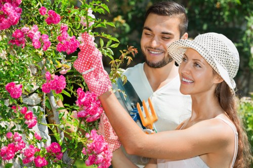 A man and woman engaging in gardening in a lush outdoor space, with the woman tending to vibrant pink flowering bushes while wearing a white dotted sun hat and pink gardening gloves. The man stands close by, smiling and holding a small gardening tool, both focused on the plants. The garden features a maintained lawn with dense green foliage and a wooden fence partially visible behind the flowering shrubs. The scene is set outdoors in natural daylight, with a backdrop of trees and greenery, indicating a well-kept residential garden ideal for outdoor maintenance services offered by landscapershounslow.co.uk, situated near the Hounslow area. The garden environment emphasizes careful plant care, pruning, and landscaping activities typical of professional gardening services in West London.