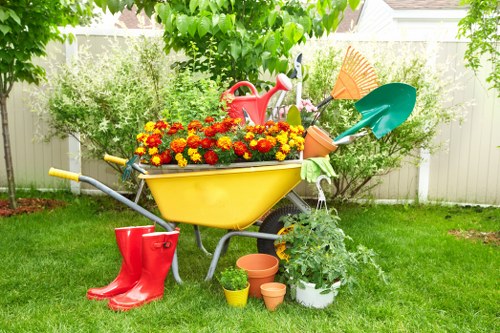 A bright, neatly maintained backyard garden with a lush green lawn and a white picket fence in the background. In the center, there is a yellow wheelbarrow filled with vibrant red and yellow flowering plants, positioned on the grass. Surrounding the wheelbarrow are various gardening tools and equipment, including a red watering can placed on top of the plants, orange and green garden trowels stuck into the soil, and a couple of terracotta plant pots placed on the ground nearby. To the left, a pair of red rubber boots rest on the grass, while a potted plant with green foliage is situated to the right, adding to the garden's natural aesthetics. The scene is illuminated by soft daylight, suggesting a mild, clear weather condition suitable for outdoor gardening activities. This setup indicates ongoing or recent gardening work in a well-kept suburban garden in Hounslow, aligning with landscaping and gardening services offered by companies like Landscapers Hounslow.