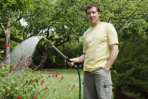 A man in a pale yellow t-shirt and grey cargo shorts is standing in a well-maintained garden in Hounslow, watering a flower bed with small red flowers using a garden hose. The garden features a lush, green lawn with dense shrubbery and canopies of leafy trees in the background, providing shade and a natural setting. The surface of the lawn is even and healthy, with a mix of grass and soil visible near the flower bed borders. The garden appears to be outdoors, in a residential or landscaped outdoor space, with clear weather conditions suggesting a sunny day. The scene reflects typical outdoor maintenance activities that Landsacpers Hounslow might undertake to keep garden areas vibrant, tidy, and inviting, supporting local gardening and landscaping services in the Hounslow area.