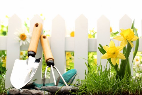 The image depicts a neatly maintained garden area in a residential outdoor space, featuring a small patch of vibrant green grass with dense, even growth and natural texture, bordered by a white picket fence that separates the garden from the background. Prominently positioned on the grass are gardening tools, including a small hand trowel and a hand rake, both with wooden handles and metal working parts, indicating recent or ongoing planting or soil cultivation. To the right, a cluster of bright yellow daffodil flowers in full bloom adds colour and natural interest, with several green leaves extending upward from their stems. In the background, the white fence is partially obscured by soft, natural daylight, suggesting a clear, sunny day with mild weather conditions typical of spring or early summer. The overall environment combines well-maintained lawn areas, flowering plants, and garden tools, illustrating an outdoor space managed by professional gardening services such as those provided by Landscapers Hounslow, aligned with their expertise in planting and garden maintenance in the Hounslow area, UK. This scene captures a tidy, inviting garden setting that highlights horticultural care and outdoor landscaping potential.