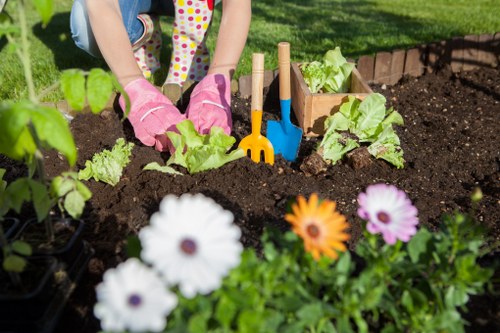 The image depicts a person's hands wearing pink gardening gloves actively planting young lettuce or similar leafy greens in a well-maintained garden bed with dark, moist soil. The garden bed is bordered by wooden planks and contains a variety of small plants, with some already established and others newly planted. In the foreground, a row of brightly coloured flowers, including white, purple, and orange blooms, adds vibrant contrast to the scene. Several small garden tools, such as a orange trowel and a blue fork, are visibly inserted into the soil, indicating ongoing planting activity. The background suggests a neat outdoor garden area, possibly part of a landscaped backyard or front garden, with lush green grass and other garden plants visible, reflecting careful outdoor maintenance typical of local gardening services like those offered by Landscapers Hounslow. The weather appears sunny and clear, providing ideal conditions for planting and garden care, aligning with garden and landscaping projects in Hounslow or nearby areas within postcode TW.