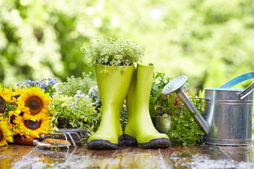 The image shows a garden scene with a rustic wooden table outdoors, featuring a pair of bright green rubber gardening boots filled with white flowering plants as a focal point. To the left of the boots, there are vibrant yellow sunflowers and other mixed flowering plants, creating a colourful and lively display. On the right side, a metal watering can with a long spout and a blue-handled garden tool are placed alongside potted greenery, suggesting ongoing garden maintenance. The surface of the table appears slightly wet, indicating recent watering or cleaning. In the background, a lush green garden with blurred trees and shrubs is visible under soft natural light, evoking a pleasant, outdoor environment suitable for gardening activities typical of local landscaping and gardening services in Hounslow. The scene captures a well-kept, cheerful outdoor space in a natural setting, highlighting garden care and landscaping elements relevant for a professional gardening service website.