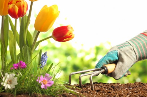 A close-up view of a person's hand wearing a gardening glove, using a hand rake to loosen rich, dark soil in a garden bed. In the foreground, there are vibrant tulips in shades of red, orange, and yellow, along with small pink, white, and purple flowers growing amidst green foliage. The garden appears well-maintained, with a variety of flowering plants and lush greenery visible in the background, suggesting a landscaped outdoor space typically found in residential gardens within Hounslow. The natural lighting indicates a bright and possibly partly cloudy day, suitable for gardening activities, supporting professional garden maintenance and landscaping services such as those offered by Landscapers Hounslow.