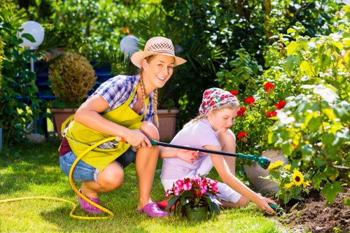 The image shows a woman and a young girl gardening together in a lush, well-maintained backyard garden. The woman, wearing a straw hat, a plaid shirt, and a yellow apron, is smiling as she assists the girl with planting or tending to flowering plants. The girl, dressed in a light-colored t-shirt and a floral headscarf, is focused on her task while using her hand to carefully work in the soil near vibrant red, yellow, and pink flowers. The garden features a neatly manicured lawn with dense greenery and various shrubs along the background, including tall trees and decorative hedges. There are patio lanterns hanging from a string above, suggesting a welcoming outdoor space for leisure. The sunny weather highlights the natural colors of the plants and the cheerful, active scene tied to outdoor gardening and lawn care services offered by Landscapers Hounslow, suitable for landscaping or garden maintenance projects in Hounslow or the surrounding areas near postcode TW or TW3.