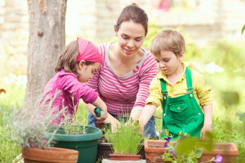 A woman with dark hair tied back, wearing a pink and white striped long-sleeve shirt, is engaging with two young children in a garden setting. One child, dressed in a pink jacket and matching pink headband, is tending to a potted plant, while the other child, wearing a yellow top and green overalls, is observing nearby. They are surrounded by various garden pots and planters containing an assortment of green leafy plants and blooming flowers, set against a backdrop of a tree trunk, blurred greenery, and a stone wall. The scene takes place outdoors on a bright, sunny day with natural light illuminating the garden area, highlighting fresh, vibrant garden life typical of a landscaped outdoor space in Hounslow or nearby London suburbs, reflecting the gardening services offered by Landscapers Hounslow.