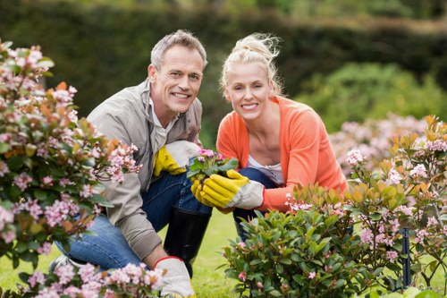 A man and a woman are gardening in a well-maintained front garden area in Hounslow, with a background of lush greenery and trees. They are kneeling among flowering bushes with pink and light purple blossoms, actively planting or tending to the plants. The man has short gray hair and is wearing a light gray shirt, while the woman has blonde hair tied back and is dressed in an orange top with gardening gloves on. The garden features neatly arranged flowering shrubs, a lush green lawn, and soil beds, all under natural daylight with clear weather. The scene captures a moment of outdoor maintenance and gardening care, highlighting the family-friendly outdoor space that could be part of professional landscaping or gardening services in Hounslow provided by Landscapers Hounslow, emphasizing a thriving, inviting garden environment.